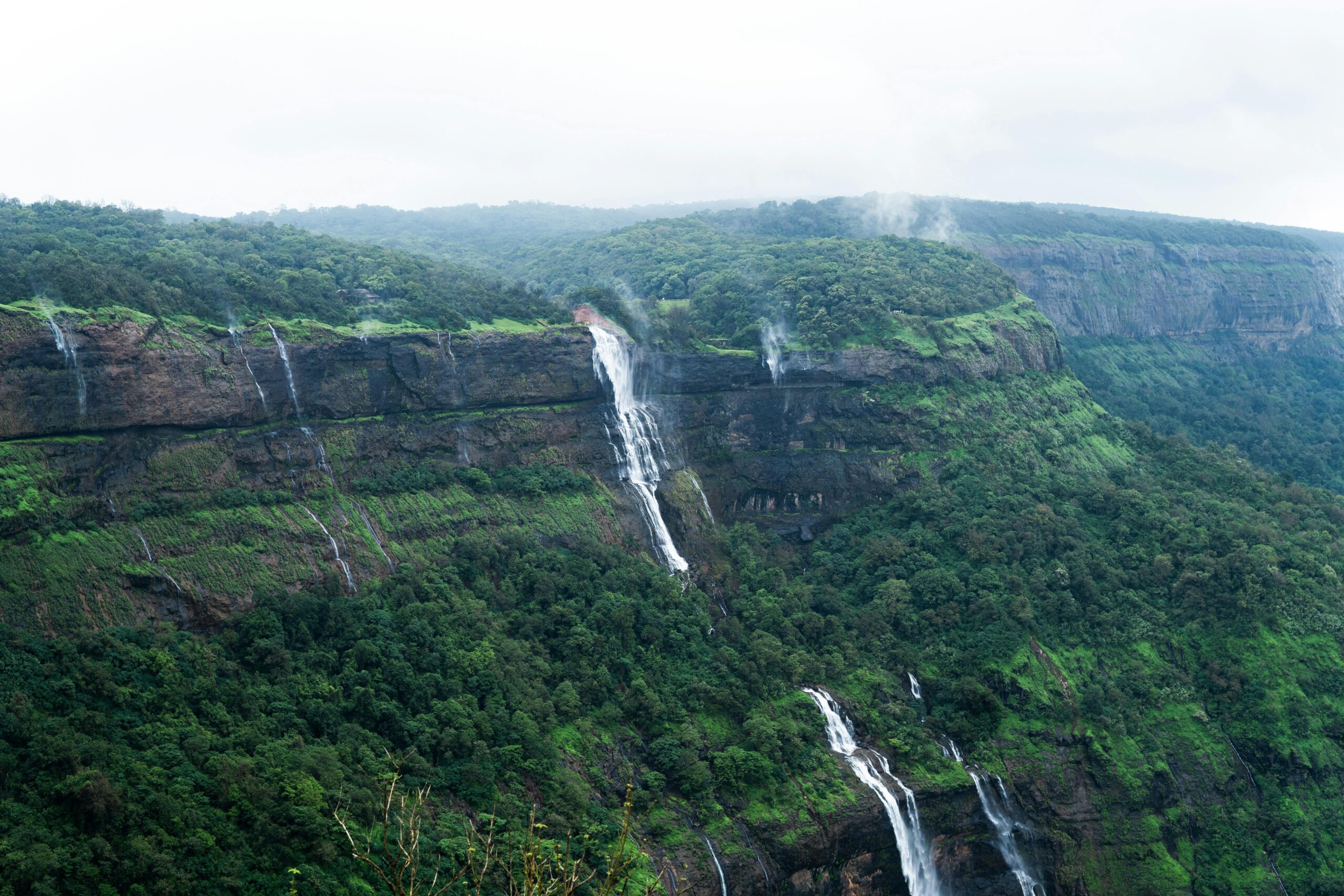 Captivating view of cascading waterfalls on a forested cliff in a tropical valley, enveloped in mist and greenery.